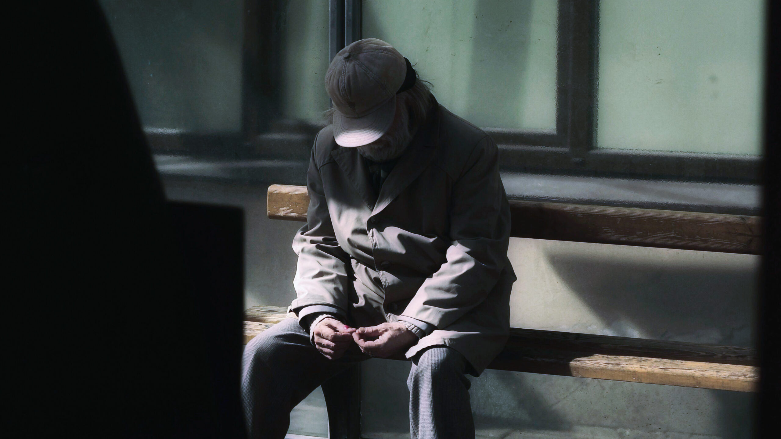 elderly man sitting on a bench alone, with dark shadows being cast on him. ageing population needs our help.