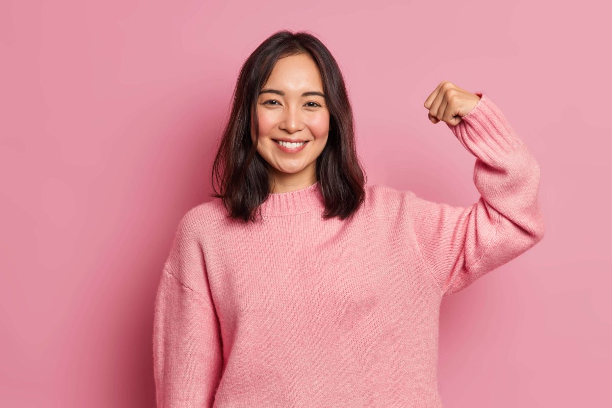 Powerful cheerful brunette woman raises arm and shows muscle demonstrates her strength looks confident at camera smiles gently wears casual long sleeved jumper isolated over pink background.